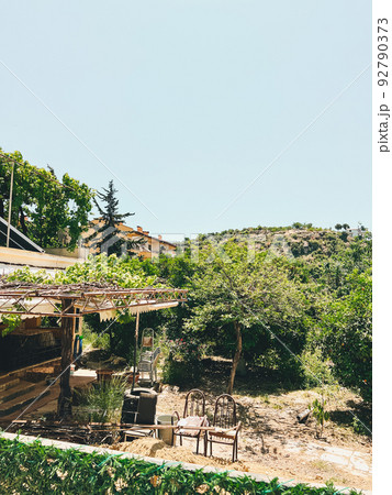Old abandoned mediterranean farm house surrounded by trees. Sun lit shabby farmhouse with old fashioned wooden furniture chairs in the garden 92790373