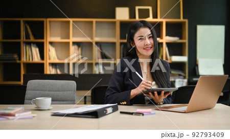 Smiling asian female manager using digital tablet and laptop while sitting in contemporary office Smiling asian female manager using digital tablet and laptop while sitting in contemporary office 92793409