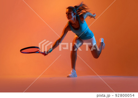 Dynamic portrait of woman, professional tennis player training isolated over orange studio background in neon light 92800054
