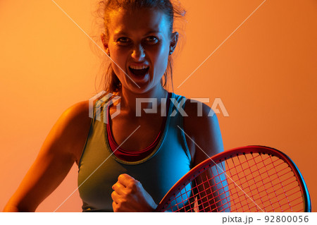 Portrait of emotive woman, professional tennis player posing isolated over orange studio background in neon light 92800056