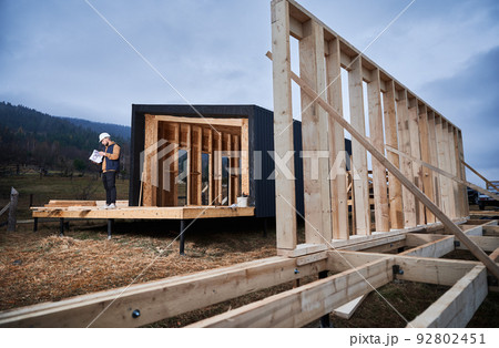 Male architect building wooden frame house in the Scandinavian style barnhouse. Man builder standing on construction site in safety helmet, with construction documentation inspecting quality of work. 92802451