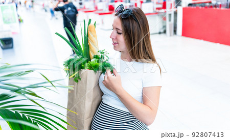 Pregnant food bag supermarket. Pregnancy woman with healthy lettuce salad leaves, fresh tomato in market food bag on grocery supermarket background. Everyday shopping concept. Pregnant food bag supermarket. Pregnancy woman with healthy lettuce salad leaves, fresh tomato in market food bag on grocery supermarket background. Everyday shopping concept. 92807413