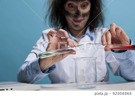 Close up of mad foolish scientist testing experimental serum by adding unknown substance while on laboratory space. Crazy chemist with messy hair and dirty face mixing unstable chemical compounds. 92808868
