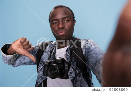 African american photography enthusiast with DSLR device showing thumbs down hand gesture at camera while standing on blue background. Photographer with photo device giving disapprove finger symbol. African american photography enthusiast with DSLR device showing thumbs down hand gesture at camera while standing on blue background. Photographer with photo device giving disapprove finger symbol. 92808869