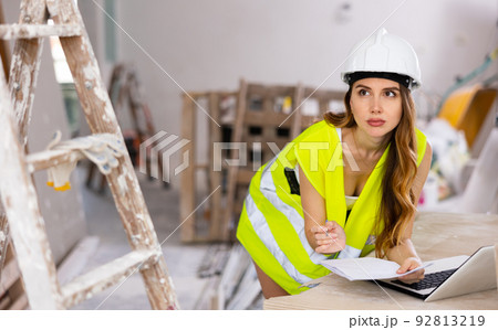 Female foreman in a protective helmet and a yellow vest checks the execution of repair work using laptop Female foreman in a protective helmet and a yellow vest checks the execution of repair work using laptop 92813219