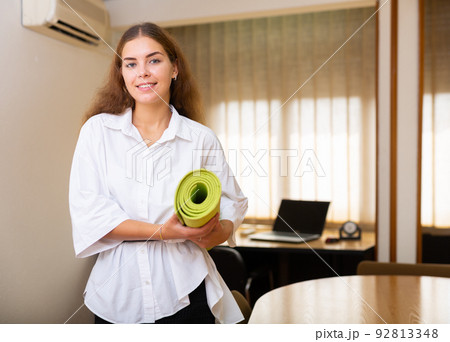 Cheerful young female entrepreneur standing with yoga matt in office 92813348