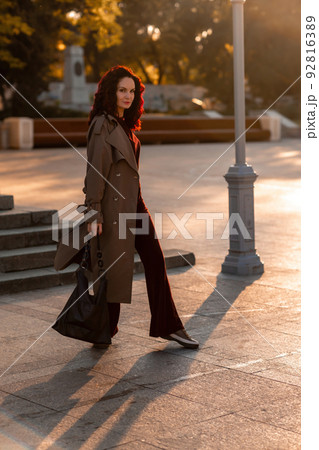 Outdoor fashion portrait of young elegant fashionable brunette woman, model in stylish hat, choker and light raincoat posing at sunset in European city. Outdoor fashion portrait of young elegant fashionable brunette woman, model in stylish hat, choker and light raincoat posing at sunset in European city. 92816389