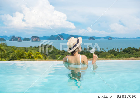 Happy woman in white swimsuit drinking wine in luxury swimming pool hotel against beautiful view, young female with hat enjoy in tropical resort. Relaxing, summer travel, holiday and vacation concept 92817090