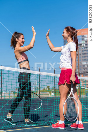 Two Caucasian women in sportswear greeting before a tennis match on an outdoor court. Players give a high five before the game. 92820055