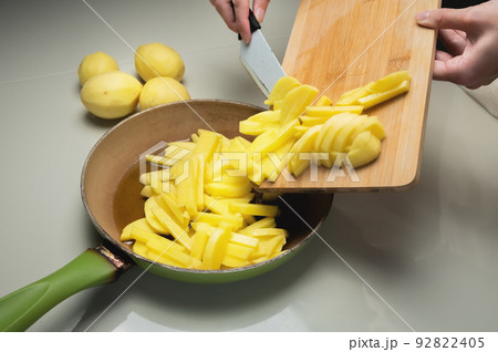 Raw potato slices on the background of a wooden cutting board with a knife, prepared and cut for frying. Placed in a frying pan by female hands Raw potato slices on the background of a wooden cutting board with a knife, prepared and cut for frying. Placed in a frying pan by female hands 92822405