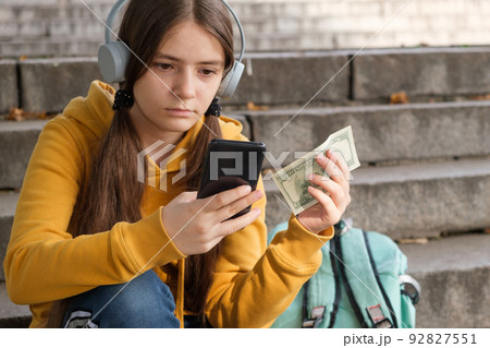 The teenager holds dollar bills and looks at the smartphone screen. The teenager holds dollar bills and looks at the smartphone screen. 92827551