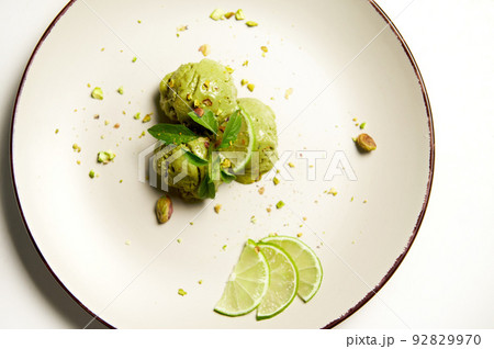 Top view. Food still life composition of dairy free, raw vegan pistachio ice cream sorbet balls, decorated with sprinkled nuts, lemon basil leaves and slices of lime on a plate, over white background Top view. Food still life composition of dairy free, raw vegan pistachio ice cream sorbet balls, decorated with sprinkled nuts, lemon basil leaves and slices of lime on a plate, over white background 92829970