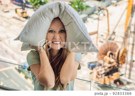 A young woman by the window annoyed by the building works outside covered my ears with a pillow. Noise concept 92833366