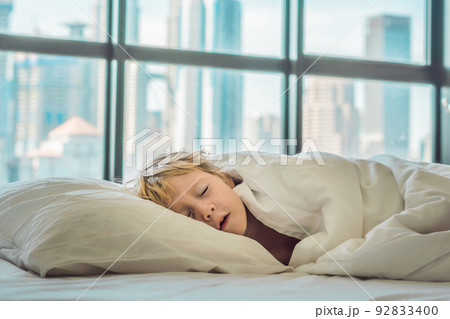 Smiling boy in bed waking up in a big city in the background of skyscrapers. Children's health in a big city concept Smiling boy in bed waking up in a big city in the background of skyscrapers. Children's health in a big city concept 92833400