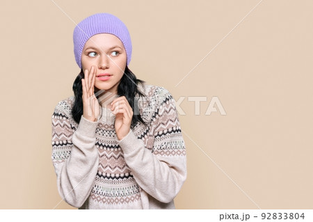Teen girl in winter sweater hat telling secret rumors, on brown background Teen girl in winter sweater hat telling secret rumors, on brown background 92833804