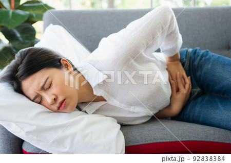 Unhappy Asian woman sitting on the sofa and holding on stomach suffering. Abdominal pain that comes from menstruation, diarrhea, or indigestion. Sickness and healthcare concept 92838384