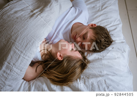 Happy young couple lie in bed and hug each other on white bed linen, look Happy young couple lie in bed and hug each other on white bed linen, look 92844505