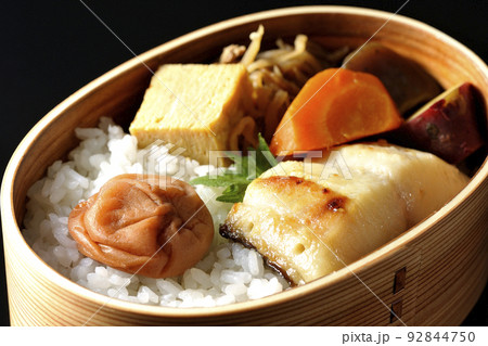 大きめの梅干しがご飯の上に乗っていて玉子焼や煮物なども入っている銀だらの西京焼き弁当を黒背景で撮影 92844750