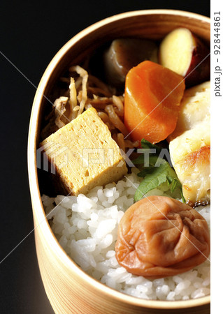 大きめの梅干しがご飯の上に乗っていて玉子焼や煮物なども入っている銀だらの西京焼き弁当を黒背景で撮影 92844861