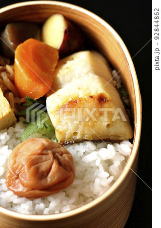 大きめの梅干しがご飯の上に乗っていて玉子焼や煮物なども入っている銀だらの西京焼き弁当を黒背景で撮影 92844862