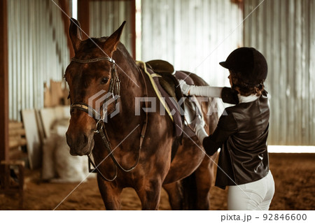 Young woman rider with brown beautiful horse, stands nearby in jockey clothes 92846600
