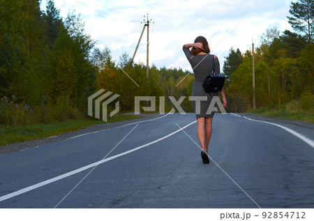 a young girl in a gray dress and black shoes with a backpack on her back, walking along an old highway against the background of a colorful forest and sunset 92854712