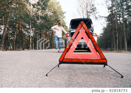 Red emergency triangle with blurred car and woman calling car mechanic in the background. Broken car on the road 92858755