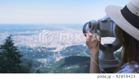 Unrecognizable woman traveler looking through viewing binoculars at summer city. Copy space. Woman in straw hat observes of blur panorama, with binoculars at observation deck. Mountain Dajti Albania 92859423