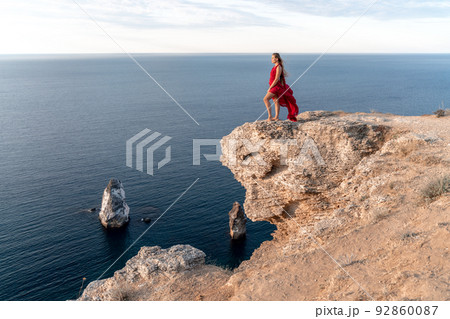 A girl with loose hair in a red dress stands on a rock rock above the sea. In the background, the sea and the rocks. The concept of travel 92860087
