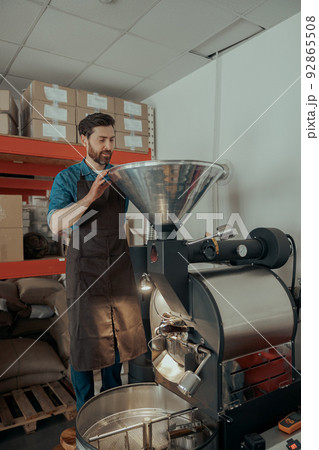 Smiling Entrepreneur pouring raw coffee beans into a modern roasting machine. Smiling Entrepreneur pouring raw coffee beans into a modern roasting machine. 92865508