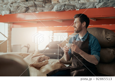 Business owner with cup of fresh coffee sitting on bags with roasted coffee beans and looking away Business owner with cup of fresh coffee sitting on bags with roasted coffee beans and looking away 92865513