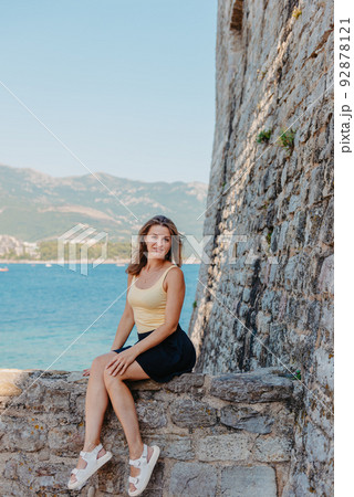 Beautiful girl sitting on a stone wall, in background is the blue sea, Budva, Montenegro. Beautiful girl sitting on a stone wall, in background is the blue sea, Budva, Montenegro. 92878121