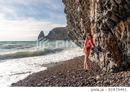 A beauty in a red swimsuit with long legs poses on a fantastic beach with huge waves against the background of mountains 92879461