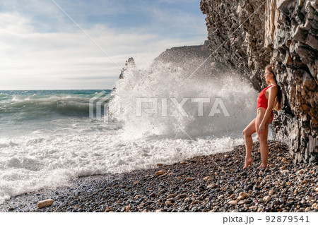 A beautiful and sexy brunette in a red swimsuit on a pebble beach, Running along the shore in the foam of the waves 92879541