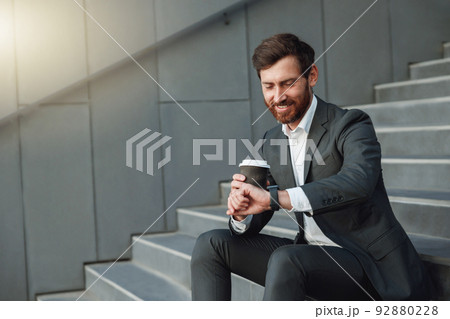 Attractive office worker in suit sitting on stairs with coffee during break and looking on his watch 92880228