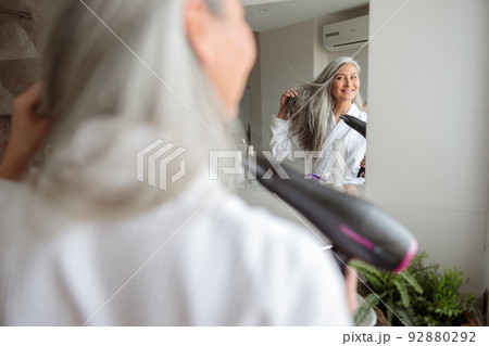 Adult woman standing and styling hair at home Adult woman standing and styling hair at home 92880292