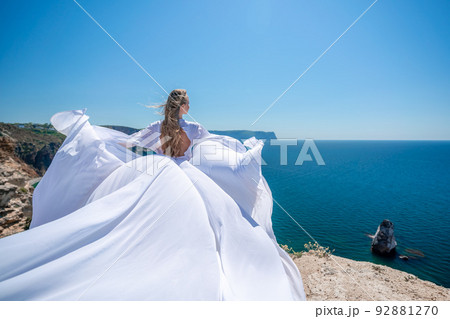 Blonde with long hair on a sunny seashore in a white flowing dress, rear view, silk fabric waving in the wind. Against the backdrop of the blue sky and mountains on the seashore. Blonde with long hair on a sunny seashore in a white flowing dress, rear view, silk fabric waving in the wind. Against the backdrop of the blue sky and mountains on the seashore. 92881270