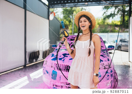 Young woman hose stands in front of a car covered in pink foam Young woman hose stands in front of a car covered in pink foam 92887011
