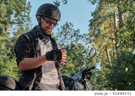 Young male biker in helmet using mobile phone and smiling sitting on a motorcycle Young male biker in helmet using mobile phone and smiling sitting on a motorcycle 92888541