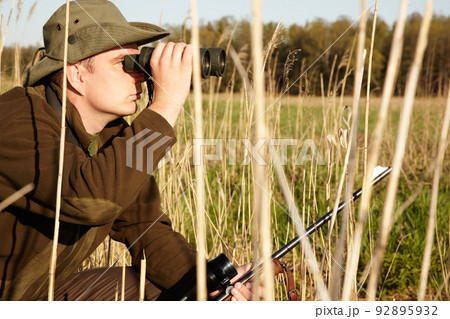 Upon closer inspection. A man in the wildlife looking through his binoculars holding his rifle. Upon closer inspection. A man in the wildlife looking through his binoculars holding his rifle. 92895932