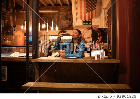 A young and happy woman eating pizza sitting alone in an empty and dark American restaurant. A female relaxing and enjoying delicious food late at night in a cosy junk food, takeaway cafe A young and happy woman eating pizza sitting alone in an empty and dark American restaurant. A female relaxing and enjoying delicious food late at night in a cosy junk food, takeaway cafe 92901829