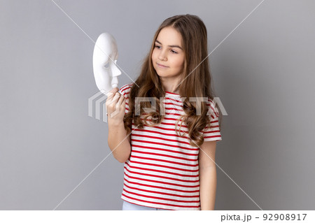 Portrait of adorable attentive dark haired little girl wearing striped T-shirt holding in hands and looking at white mask with interest. Indoor studio shot isolated on gray background. 92908917