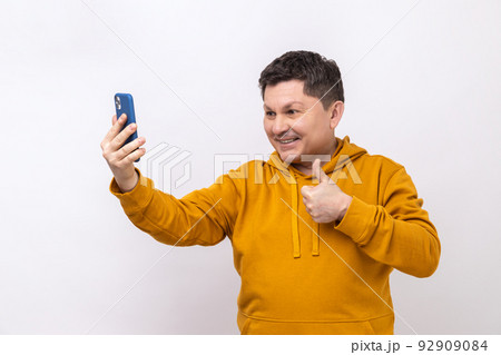 Smiling man showing thumbs up like gesture and looking at phone camera, making selfie or recording video, wearing urban style hoodie. Indoor studio shot isolated on white background. 92909084