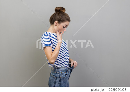 Portrait of beautiful surprised woman wearing striped T-shirt looking at her slim waist in big trousers with open mouth, successful weight loss. Indoor studio shot isolated on gray background. 92909103