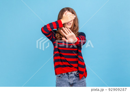 I dont want to see it. Portrait of woman wearing striped casual style sweater covering eyes with hand and gesturing stop, feeling ashamed or scared. Indoor studio shot isolated on blue background. 92909287