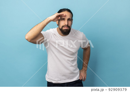 Portrait of curious man with beard wearing T-shirt keeping palm over head and looking attentively far away, peering with expectation at long distance. Indoor studio shot isolated on blue background. Portrait of curious man with beard wearing T-shirt keeping palm over head and looking attentively far away, peering with expectation at long distance. Indoor studio shot isolated on blue background. 92909379