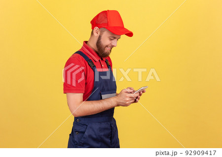 Side view of smiling worker man typing on mobile phone, using cellphone messenger to accept online order as delivery, repair and maintenance services. Indoor studio shot isolated on yellow background. 92909417