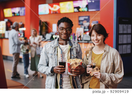 Portrait of young Black man and Caucasian woman wearing casual clothes having date at cinema holding popcorn, drinks and tickets smiling at camera 92909419
