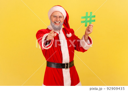 Positive elderly man with gray beard wearing santa claus costume standing with green hashtag in hands, pointing to camera with toothy smile. Indoor studio shot isolated on yellow background. 92909435