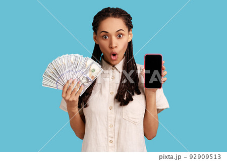 Attractive shocked woman with black dreadlocks holding fan of dollar banknotes and smart phone with empty screen, making money online. Indoor studio shot isolated on blue background. 92909513